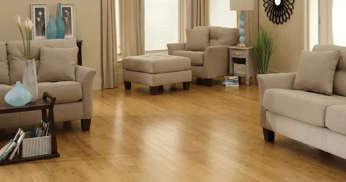 A living room with professionally installed bamboo flooring, beige sofas, a matching ottoman, a side table with a blue lamp, vases, framed pictures, and large windows with blinds and curtains in Staten Island, NY